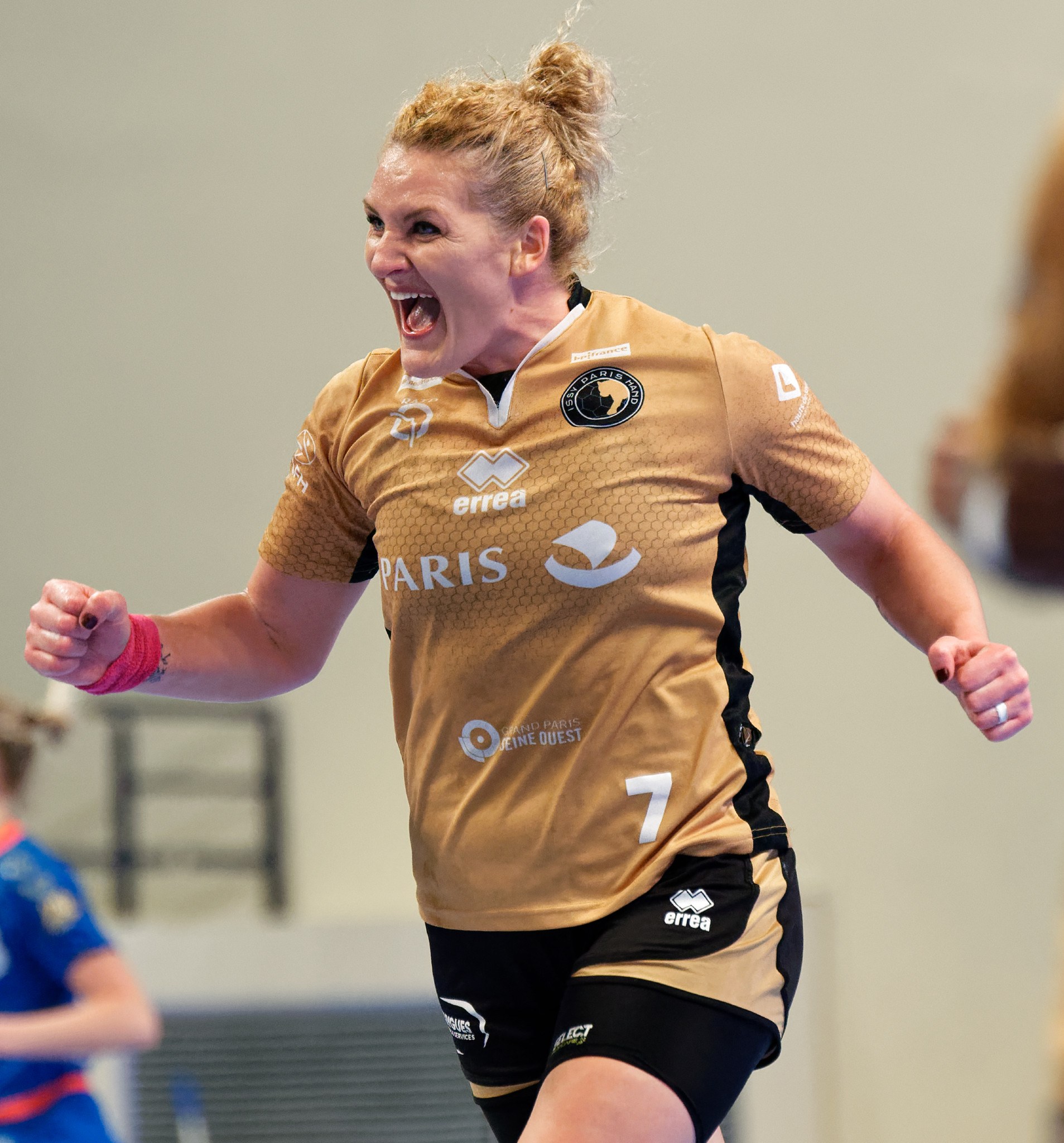 Eine Frau in brauner Bluse und schwarzen Shorts, die auf einem Futsalplatz läuft und feiert, mit zwei anderen Frauen im Hintergrund.
