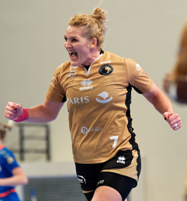 Eine Frau in brauner Bluse und schwarzen Shorts, die auf einem Futsalplatz läuft und feiert, mit zwei anderen Frauen im Hintergrund.