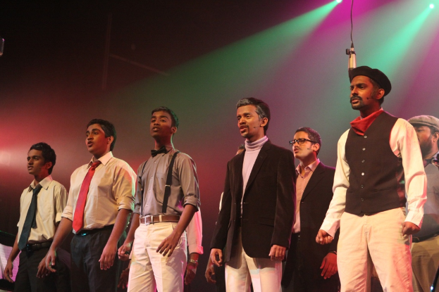 Eine Gruppe von Männern steht auf einer Bühne und spielt ein Drama, während grüne Spotlights hinter ihnen leuchten.