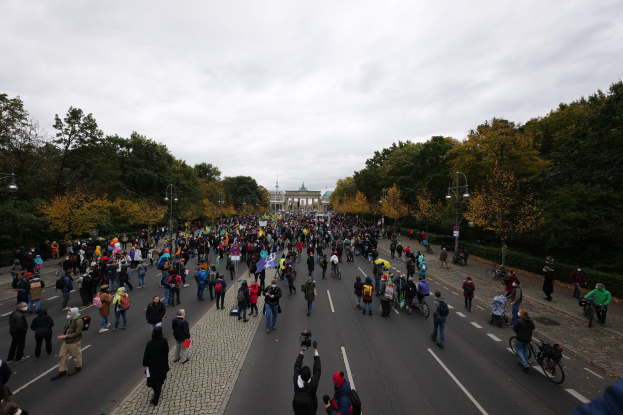 Eine große Gruppe von Menschen, die eine von Bäumen gesäumte Straße in Berlin entlanggeht, einige halten Kameras, mit Gebäuden und einem klaren Himmel im Hintergrund.