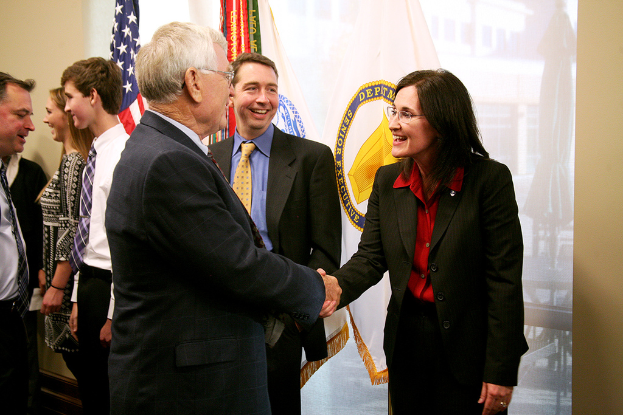Ein Mann und eine Frau in Blazern und Hemden geben sich die Hand und lächeln sich an, während ein Mann in Blazer, Krawatte und Hemd neben ihnen steht; im Hintergrund unterhalten sich Menschen und lächeln mit Fahnen an der Wand.