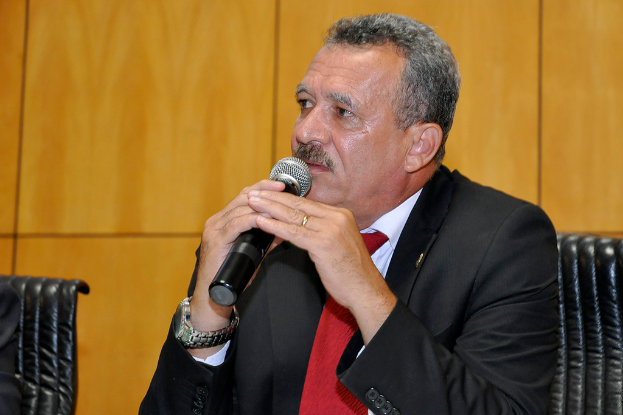 Ein Mann in Anzug und Uhr sitzt auf einem Stuhl und hält ein Mikrofon in der Hand, neben ihm steht ein leerer Stuhl und eine hölzerne Wand im Hintergrund.