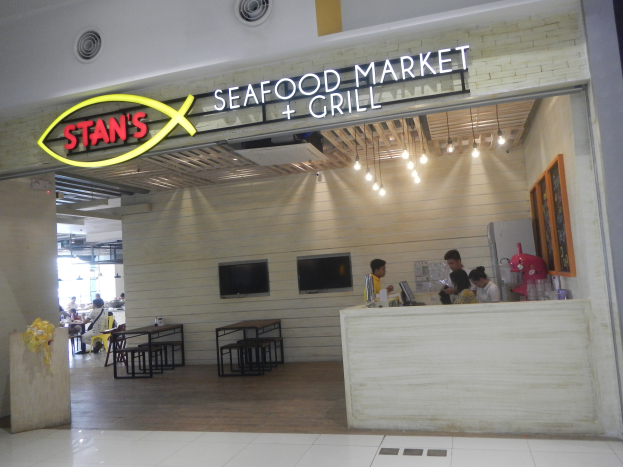 Im Innern von Stans Seafood Market & Grill sitzen und stehen Menschen, auf einem Tisch sind Gegenstände und an der rechten Wand ein Foto Rahmen zu sehen, im Hintergrund Fernseher und Lampen.