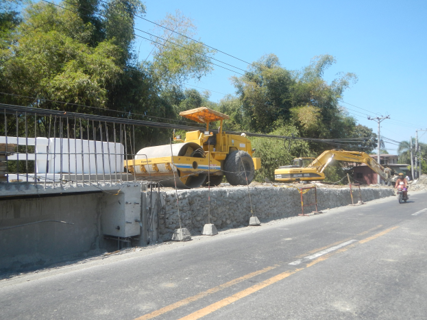 Baustelle mit einer Walze, Fahrzeugen, einem Radfahrer, einem Zaun, Steinen, einer Wand, Bäumen, Strommasten mit Drähten, einem Haus und einem bewölkten Himmel.