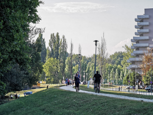 Eine Gruppe von Menschen fährt mit Fahrrädern auf einem Parkweg mit umgebender Vegetation, Straßeninfrastruktur, Fahrzeugen und Gebäuden unter einem bewölkten Himmel.