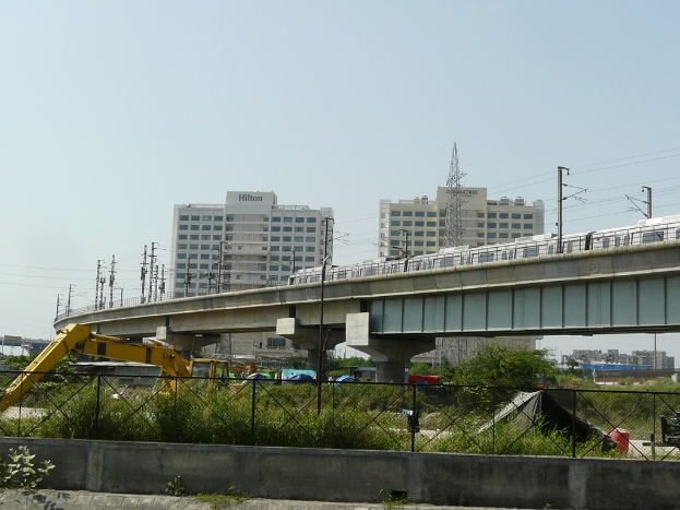 Ein klarer Himmel mit Gebäuden unten, eine Brücke mit einem darüberfahrenden U-Bahn-Zug, Strommasten, die durch Drähte verbunden sind, ein Pflugfahrzeug, Pflanzen, Bäume, ein schwarzes Zelt und ein Zaun.