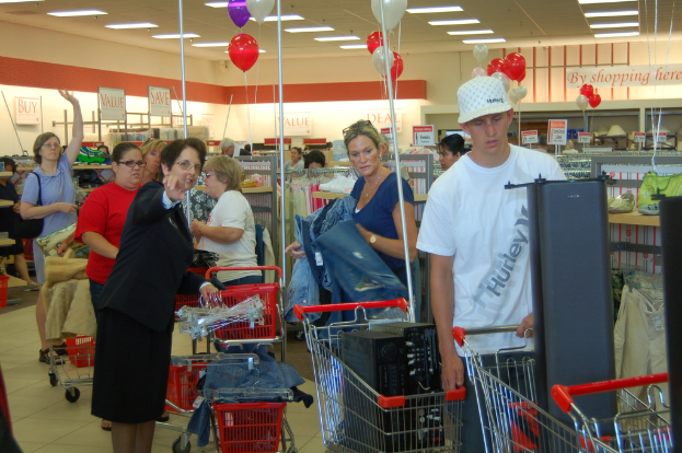 Menschen shoppen in einem Geschäft mit Einkaufswagen, die mit Waren gefüllt sind, und elektronischen Geräten auf Regalen im Hintergrund sowie Ballons und Lichter an der Decke.