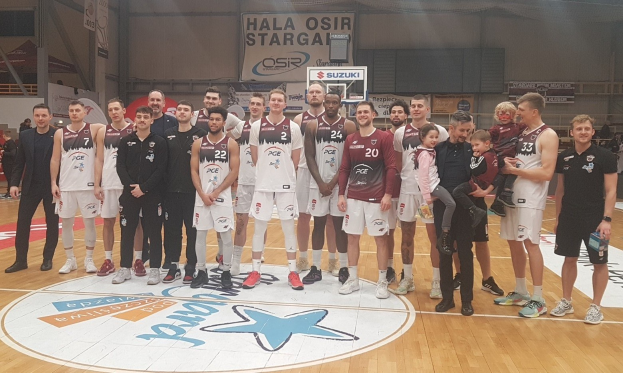 Eine Gruppe von Männern und Kindern auf einem Basketballfeld mit Schildern, die "Hala Osir Starga" lesen, umgeben von sitzenden Zuschauern, einem Basketballkorb, Metallstangen, Deckenlampen und einer Wand.