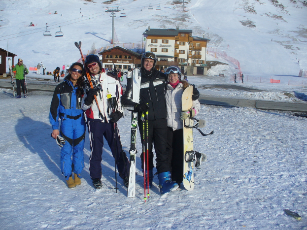 Vier Personen posen für ein Foto mit Skate-Pads und Werkzeugen, mit Gebäuden und anderen Menschen im Hintergrund, vor einer schneebedeckten Berglandschaft.