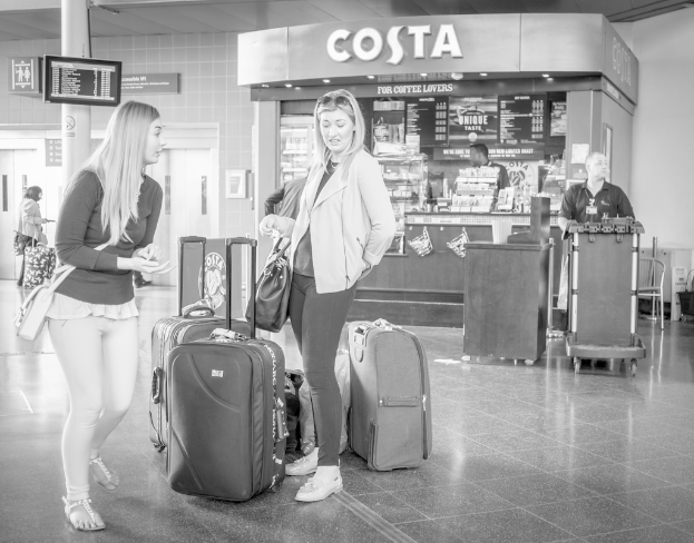 Zwei Frauen mit Gepäcktaschen vor einem Kaffeehaus in einer Umgebung, die wie ein Flughafen aussieht.