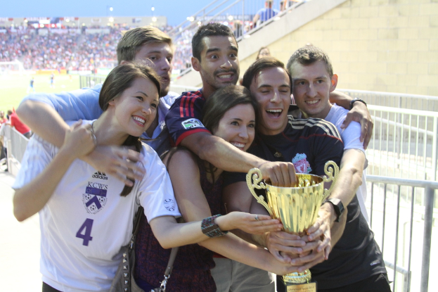 Sechs Menschen, zwei Frauen und vier Männer, halten eine Trophäe und lächeln für ein Foto vor einem Eisengeländer, mit einer Wand und einem Geländer oben rechts und Menschen und Lichtern im Hintergrund, möglicherweise in einem Stadion.