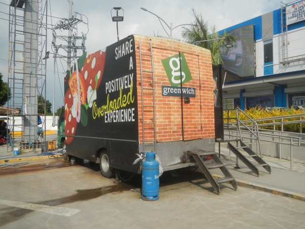 Ein großer Lastwagen steht vor einem Gebäude mit einer Tafel, auf der 'Teilen Sie eine positiv überladene Erfahrung' steht, mit Stufen, einem Zylinder auf dem Boden, Geländern, Strommasten, Laternen, Bäumen und einem bewölkten Himmel.