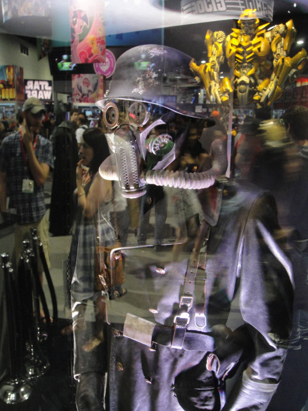 Ein Spiegelbild in einer Halle, das eine Person in Anzug und Helm in der Vordergrund, eine Gruppe von Menschen, Stangen, Poster mit Text, Bilder, ein gelbes Spielzeug und verschiedene andere Gegenstände und Lichter zeigt.