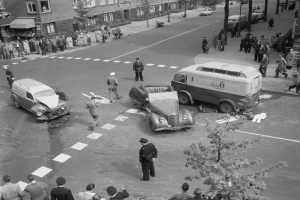 Schwarz-weiß-Szene eines Autounfalls an einer Straßenkreuzung mit mehreren Fahrzeugen, darunter ein Lieferwagen, und einer Gruppe von Zuschauern; Gebäude, Bäume, Pfosten und Texttafeln sind im Hintergrund zu sehen.