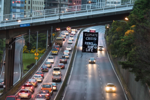 Eine belebte Stadtstraße mit starkem Verkehr, eine Brücke im Hintergrund und ein Banner mit Text in der Mitte der Straße, flankiert von Bäumen, Pfählen, Laternen, Schildern und Gebäuden auf der rechten Seite.