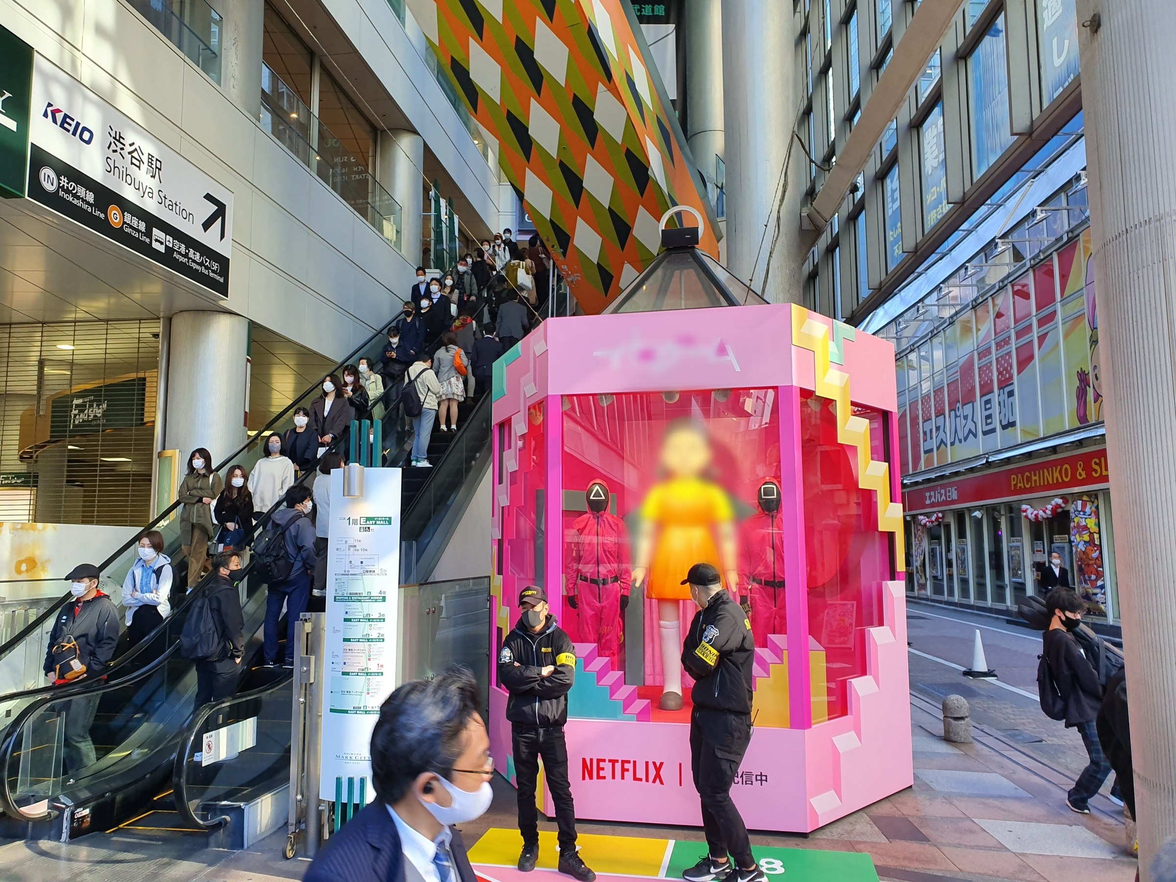 Eine Gruppe von Menschen steht vor einem gläsernen Gebäude mit Säulen und Texttafeln, einige tragen Masken, während eine Rolltreppe mit Menschen im Hintergrund zu sehen ist; eine rosafarbene Box in der Mitte zeigt Schaufensterpuppen und trägt die Aufschrift "Netflix's new Netflix booth in Tokyo."