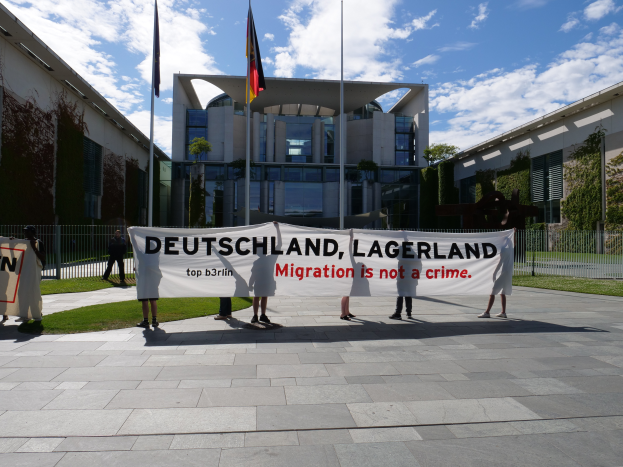 Eine Gruppe von Menschen hält ein Transparent mit der Aufschrift "Deutschland, Lagerland Migration ist kein Verbrechen" vor einem Gebäude mit Fenstern, umgeben von Gras, einem Metallzaun, Pflanzen, Bäumen und Fahnen auf Stangen, unter einem bewölkten Himmel.