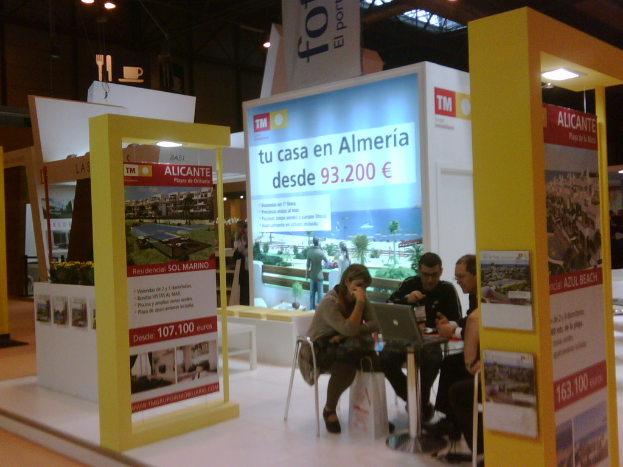 Eine Gruppe von Menschen sitzt um einen Tisch, schaut auf einen Laptop und sieht verschiedene Werbeanzeigen auf den Fleexi-Bildschirmen daneben.