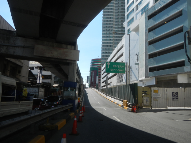 Eine Stadtstraße mit Verkehrskegeln am Straßenrand, Fahrzeuge auf der Straße, eine Brücke links, Gebäude mit Namensschildern auf beiden Seiten, Laternenpfähle und ein Himmel im Hintergrund.