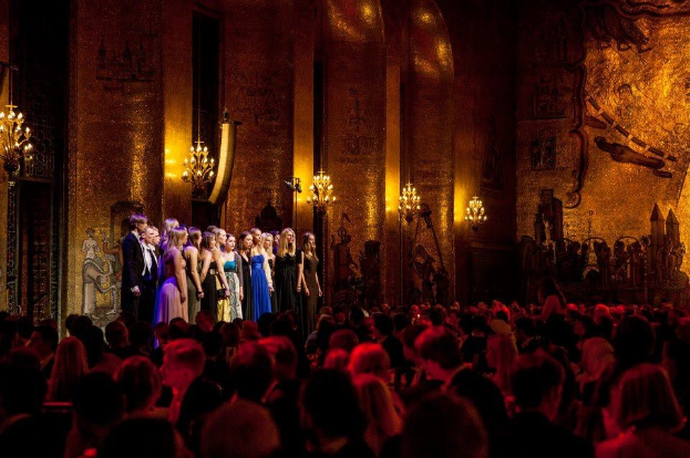 Gruppe von Menschen auf einer beleuchteten Bühne mit bemalten Wänden an der Royal Opera House, betrachtet von einem Publikum.