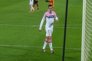 Ein Fussballspieler steht auf einem Feld neben einem Tor mit Netz auf der rechten Seite, während andere Spieler im Hintergrund sichtbar sind.