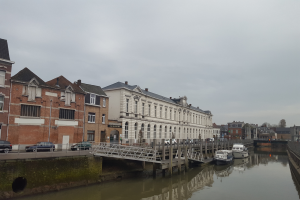 Ein Kanal mit anlegten Booten, eine Brücke mit Geländern, Fahrzeuge auf der Straße, Gebäude mit Fenstern, Laternenpfähle, Bäume und ein bewölkter Himmel im Hintergrund.