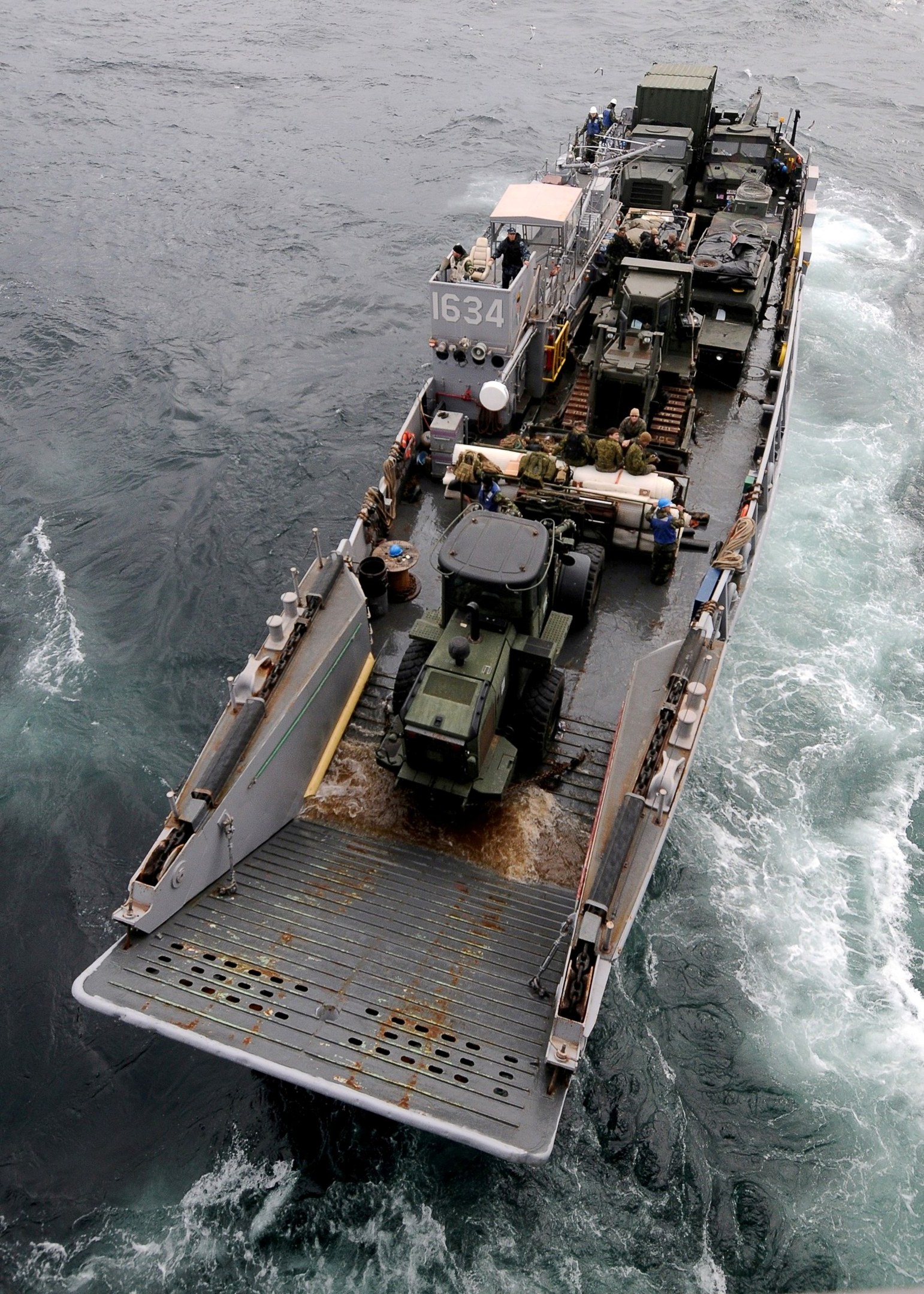 Luftaufnahme eines großen Bootes auf dem Meer mit einem Militärfahrzeug darauf, umgeben von Menschen und Equipment, mit einem Wasserzeichen unten.