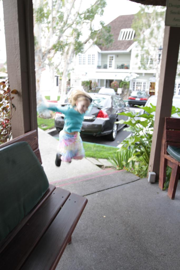 Ein Mädchen springt vor einer Bank mit Autos, Gebäuden und Bäumen im Hintergrund.