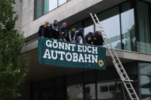 Eine Gruppe von Menschen steht auf dem Dach eines Gebäudes und hält eine Fahne, mit einem Baum links und Glasfenstern im Hintergrund, wahrscheinlich protestieren sie im Support der deutschen Regierung.