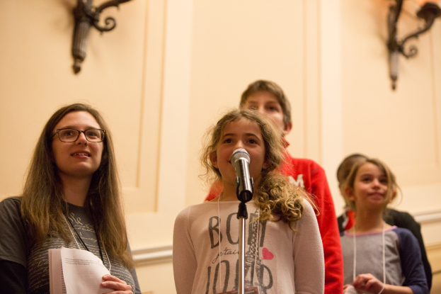 Eine Gruppe junger Mädchen steht nebeneinander vor einem Mikrofon, eine hält ein Blatt Papier in der Hand, im Hintergrund sind Wandlampen zu sehen, die eine gemütliche Atmosphäre schaffen.