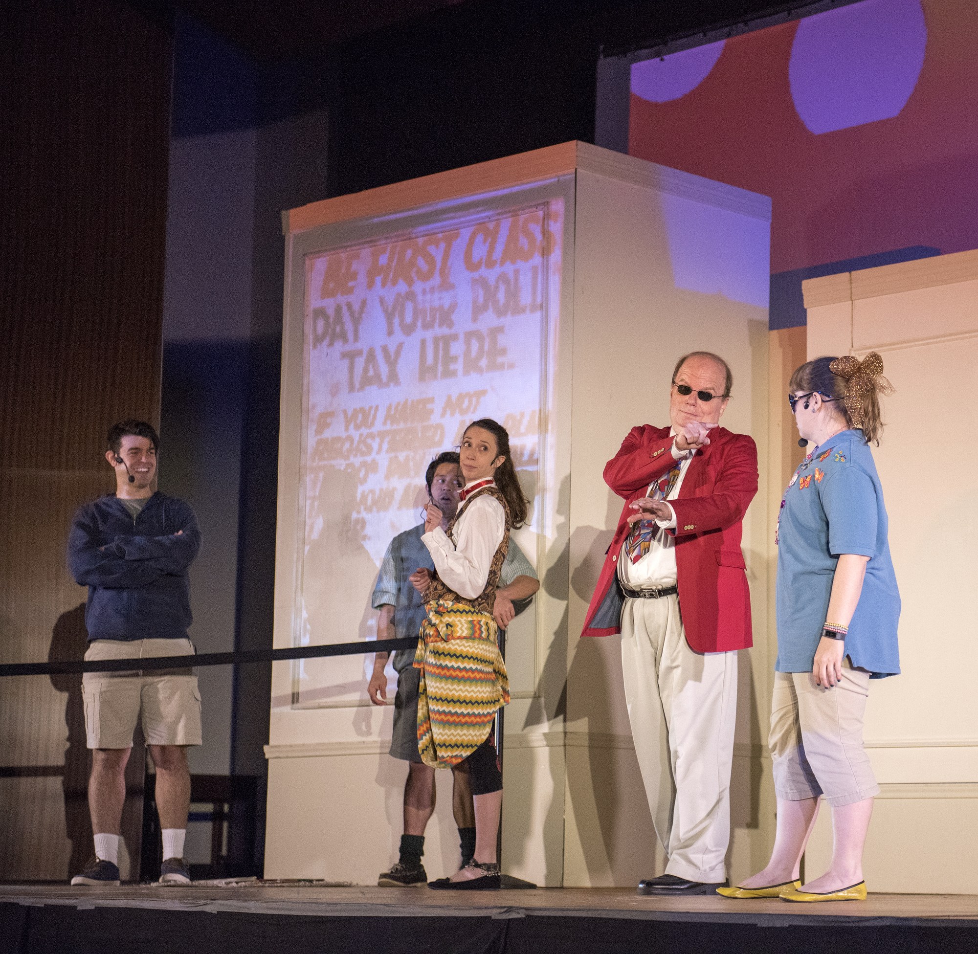 Eine Gruppe von Menschen auf einer Bühne mit einem Mann, der ein Mikrofon hält, ein Display mit der Aufschrift "Der erste Klasse-Tag Deine Steuer hier", ein Zaun, eine Wand und Deckenleuchten.