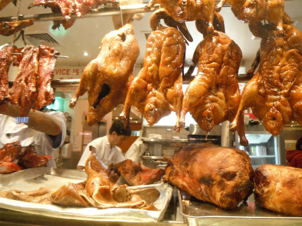 Chicken pieces hang from a rod at the top, with trays of meat below, two men standing behind the trays, racks with few items, a refrigerator, a named wall in the background, and lights on the ceiling.