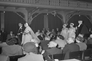 Ein Schwarz-Weiß-Foto einer Hochzeitszeremonie in einem großen Raum mit Gästen auf Stühlen und Stehenden, Sträuße, Tassen und andere Gegenstände auf Tischen sowie Vorhänge und Deckenleuchten im Hintergrund.