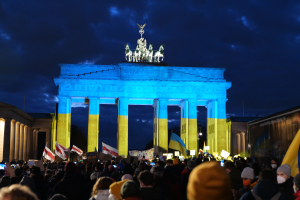 Eine Menschenmenge steht vor dem Brandenburger Tor in Berlin, Deutschland, mit Fahnen und Schildern, einem Banner auf der rechten Seite und dem Tor mit Statuen und Säulen unter einem bewölkten Himmel.