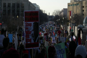 Menschen demonstrieren auf einer Straße und halten Plakate, mit Gebäuden, Bäumen, Laternenmasten und dem Himmel im Hintergrund.