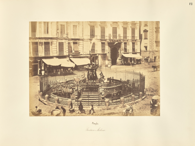 Ein altes Foto eines Stadtplatzes mit einem Springbrunnen, der von einem Metallzaun umgeben ist, Menschen, die auf Bänken stehen und sitzen, Zelte und ein Gebäude mit Fenstern im Hintergrund, mit Text am unteren Rand des Bildes.