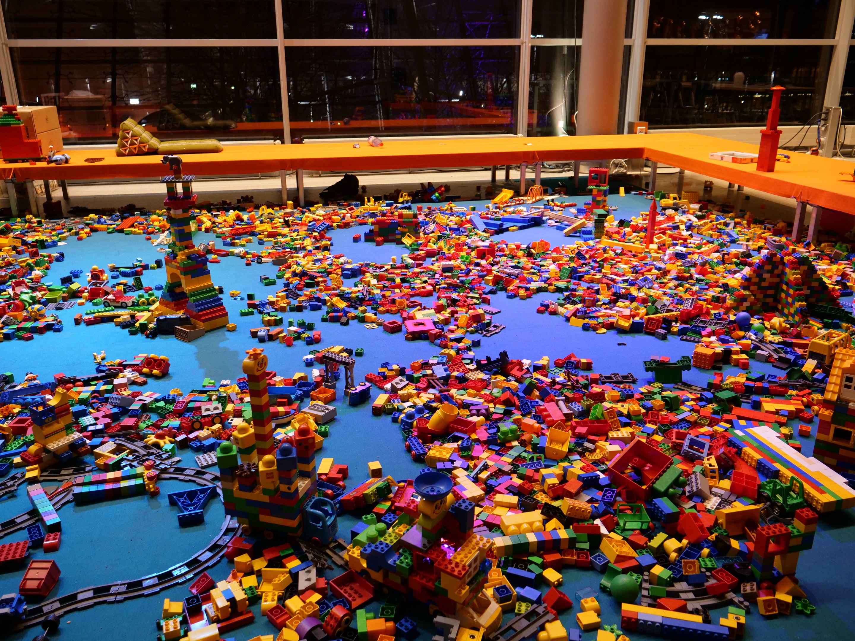 Ein großer Raum mit farbigen Lego-Blöcken, die auf dem Boden verstreut sind, umgeben von Tischen, Säulen und Glasfenstern, durch die Menschen und Ausstellungen zu sehen sind.