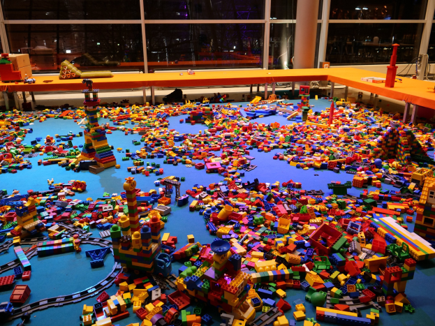 Ein großer Raum mit farbigen Lego-Blöcken, die auf dem Boden verstreut sind, umgeben von Tischen, Säulen und Glasfenstern, durch die Menschen und Ausstellungen zu sehen sind.