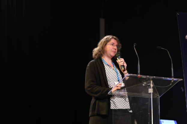 Eine Frau in einer schwarzen Jacke, die ein Mikrofon hält, mit einem dunklen Bild.