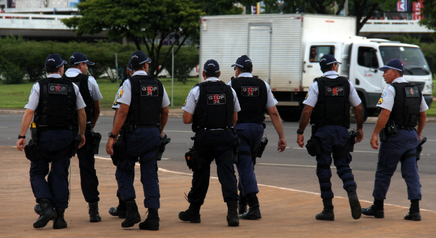 Eine Gruppe von Beamten in Uniform, einschließlich Mützen, Hemden, Schutzwesten, Hosen und Schuhen, die auf einer Straße mit einem geparkten Lastwagen auf der rechten Seite neben einem Zaun gehen; im Hintergrund sind Gebäude, Bäume, Pflanzen, Pfosten, Banner und eine Brücke in der oberen rechten Ecke zu sehen.