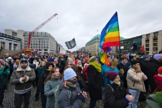 Große Gruppe von Menschen bei einer LGBTQ+-Rechtsdemo in Berlin, die Fahnen und Plakate schwingt, mit Gebäuden, einem Kran und Wolken im Hintergrund.