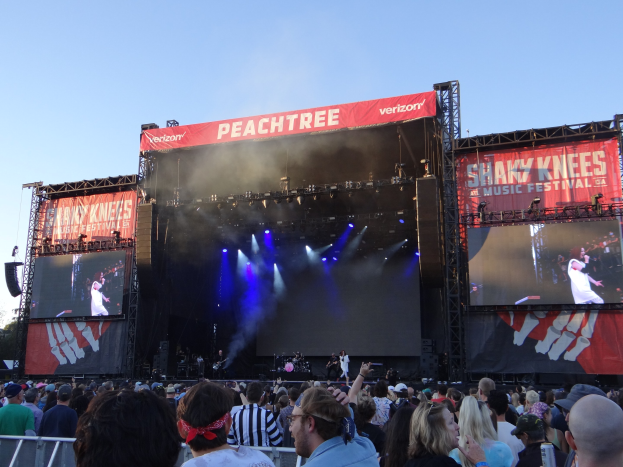 Eine große Menschenmenge steht vor einer hell erleuchteten Bühne auf dem Shaky Knees Music Festival, wo Musiker unter Schildern mit Text spielen, mit einem Geländer im Vordergrund und einem klaren blauen Himmel im Hintergrund.