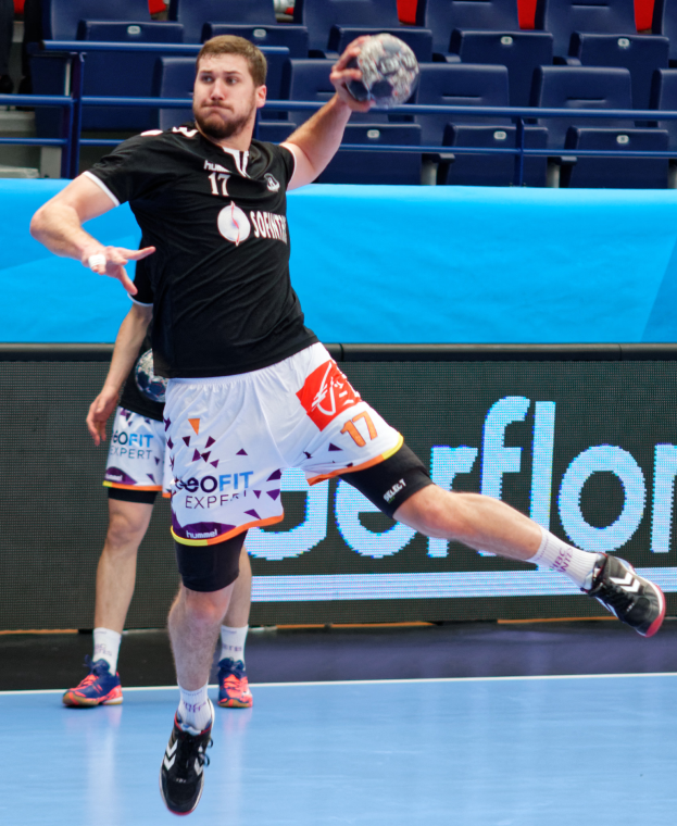 Ein Mann in schwarzem Hemd und weißen Shorts hält einen Handball und bereitet sich auf das Spiel vor, mit Zuschauern im Hintergrund einer Stadion.