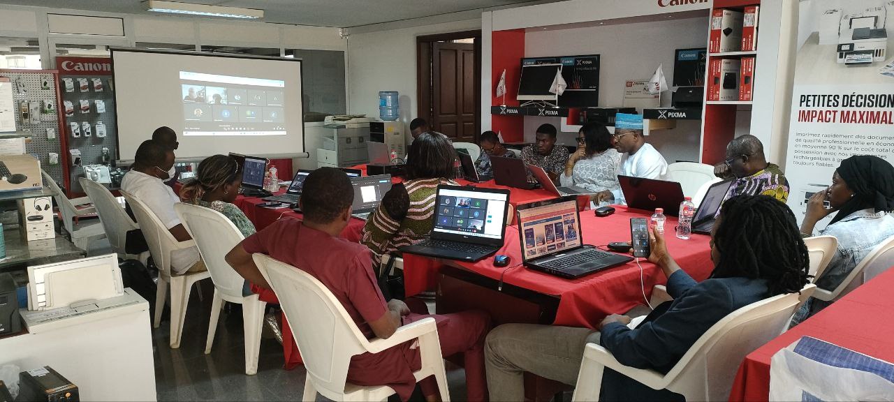 Gruppe von Menschen, die um einen Tisch mit Laptops, Flaschen und anderen Gegenständen sitzen; Banner mit Text an der Wand, Anzeigebildschirm, Tür und Deckenbeleuchtung im Hintergrund.