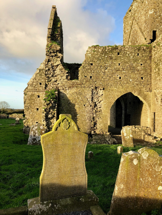 Ruinen einer alten Kirche in Irland mit einem Friedhof im Vordergrund und einer Burg im Hintergrund bei einem bewölkten Himmel, mit einem Wasserzeichen.