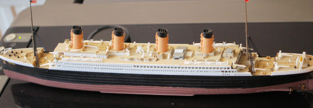 Ein creme-, weiß-, schwarz-, braun- und orangefarbenes Spielzeugschiff mit je zwei Polen und zwei Flaggen auf jeder Seite, das über einem druckerähnlichen Objekt positioniert ist.