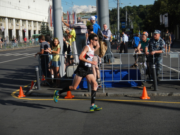 Ein Mann beim Marathonlauf auf einer Stadtstraße, umgeben von Zuschauern mit Fahrrädern, mit städtischer Infrastruktur und einem bewölkten Himmel.