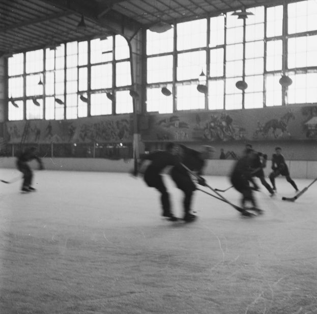 Eine Gruppe von Menschen, die Hockey auf einem Eisplatz spielen, mit einer Wand im Hintergrund, die Gemälde und Glasfenster zeigt, in Schwarz-Weiß dargestellt.