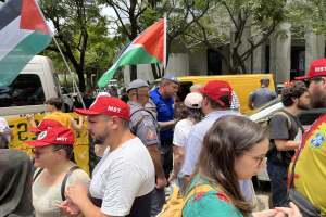 Eine Gruppe von Menschen, einige mit Mützen und Taschen bekleidet und palästinensische Flaggen tragend, steht vor einem Gebäude, während Fahrzeuge auf der Straße und Bäume im Hintergrund zu sehen sind.