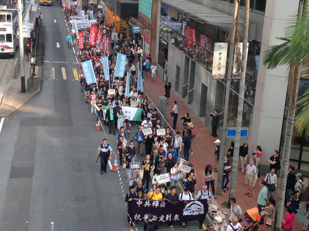 Eine große Gruppe von Menschen marschiert auf einer Straße in Hong Kong, mit Schildern und Plakaten in der Hand, während Bäume, Glasfassaden, Fahrzeuge und Schilder auf beiden Seiten zu sehen sind.